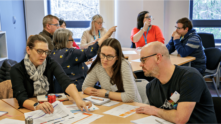 Group of secondary computer science teachers engaged in a discussion around a classroom table with papers and charts laid out, in a well-lit CPD course setting.